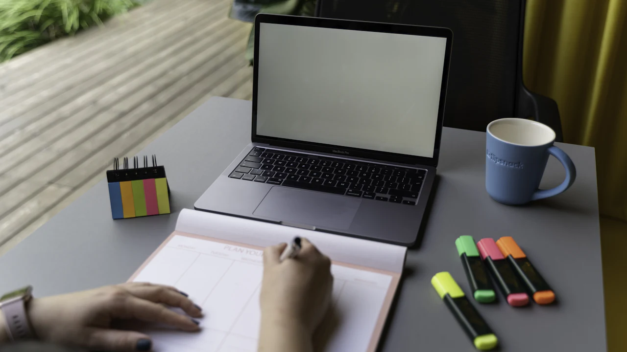 A person drafts a plan beside a laptop, calendar, mug, and markers