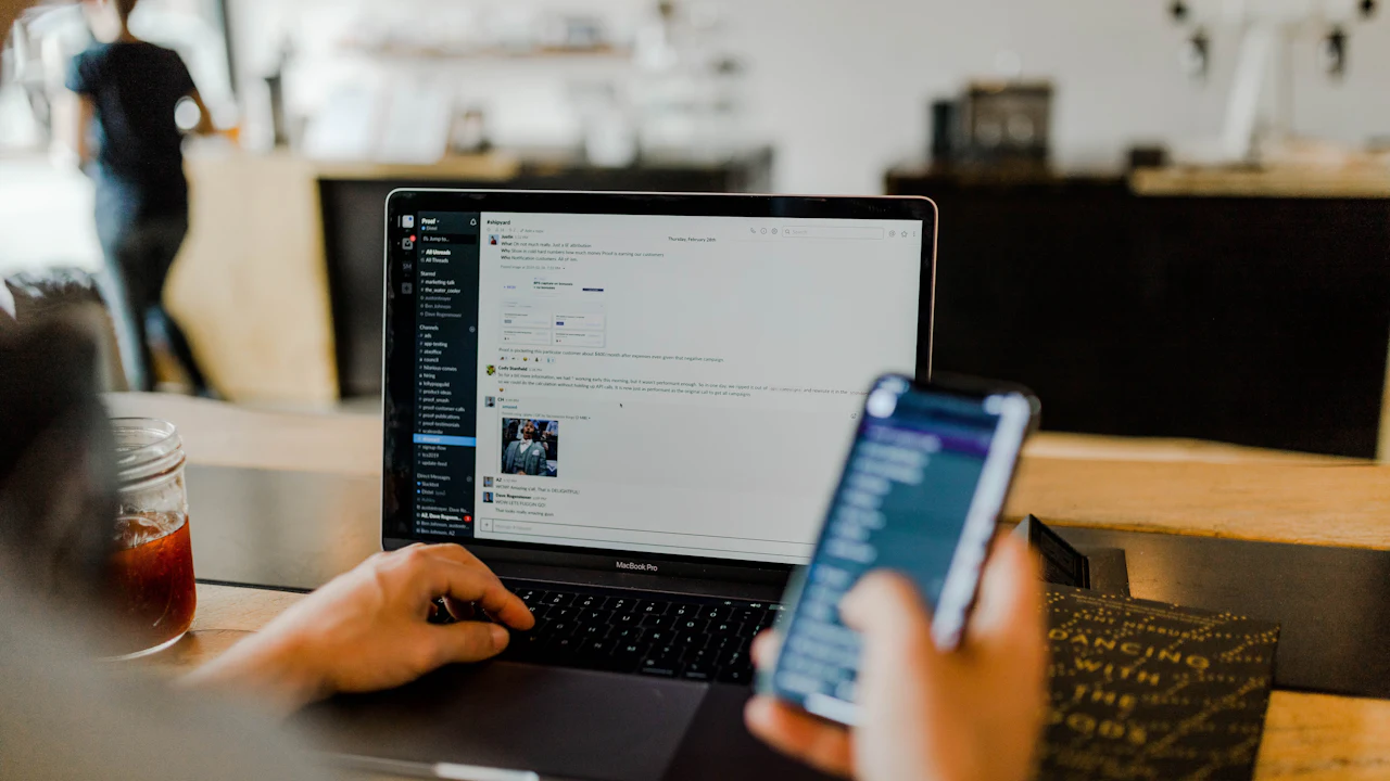 A person checks a team message on a smartphone beside a laptop